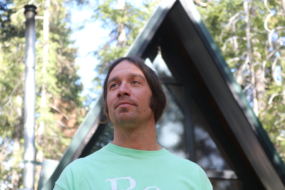 Man stands in front of his A-frame home in the Tahoe forest.